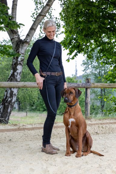 Eine Person in schwarzer Kleidung steht neben einem braunen Hund auf einem Weg.